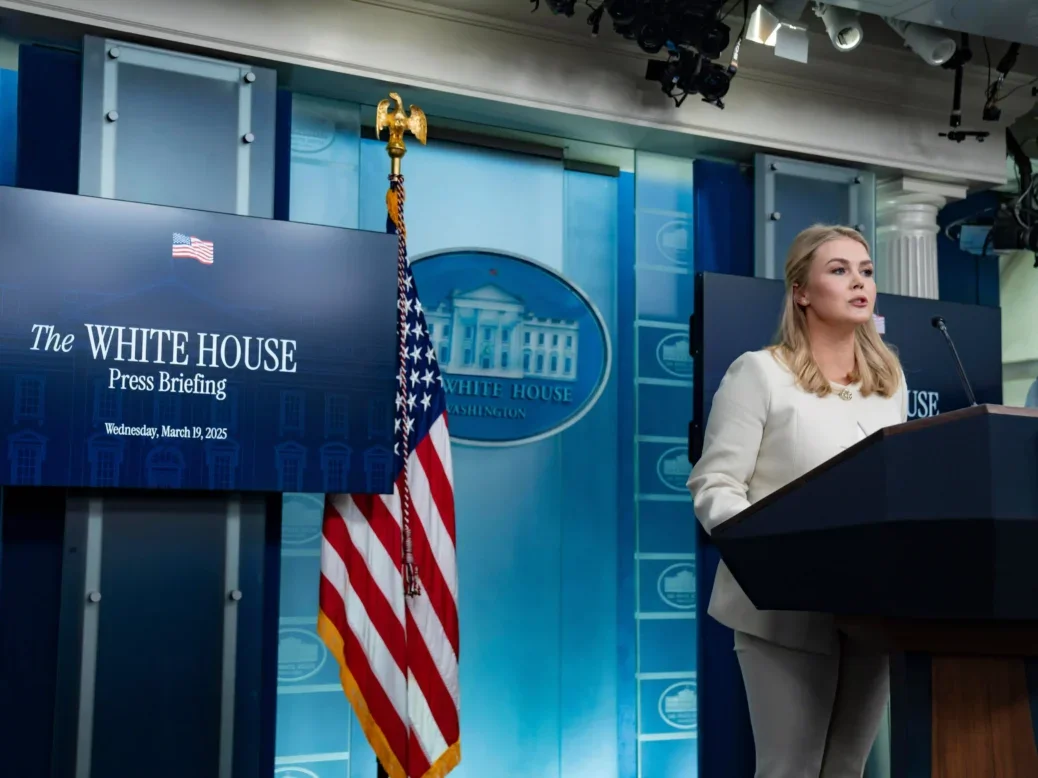 White House press secretary Karoline Leavitt conducts a media briefing. Leavitt, a young woman with blonde hair wearing a white pantsuit, is pictured mid-speech behind a podium and in front of a US flag, a screen reading "White House Press Briefing" and a seal depicting the presidential home.