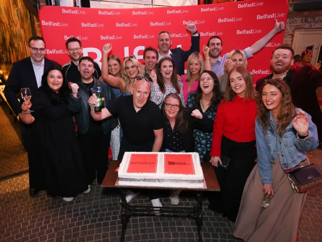 Belfast Live team pictured at event on 3 April 2025 to mark the brand's tenth anniversary. Group of about 20 people cheering, some with arms raised, in front of Belfast Live backdrop and with a Belfast Live cake